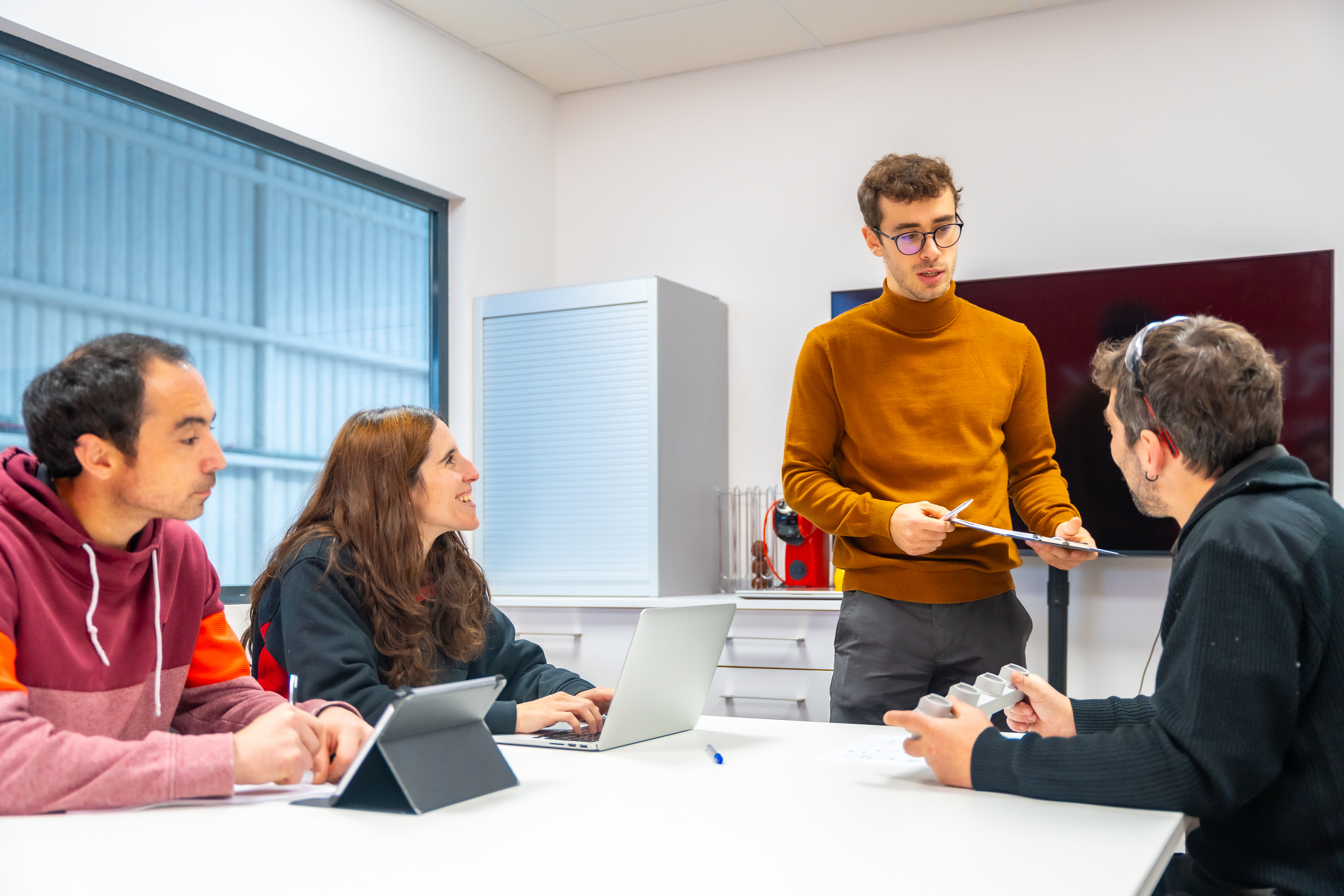 Foto de equipe em mesa de trabalho, com três pessoas sentadas e uma em pé conversando