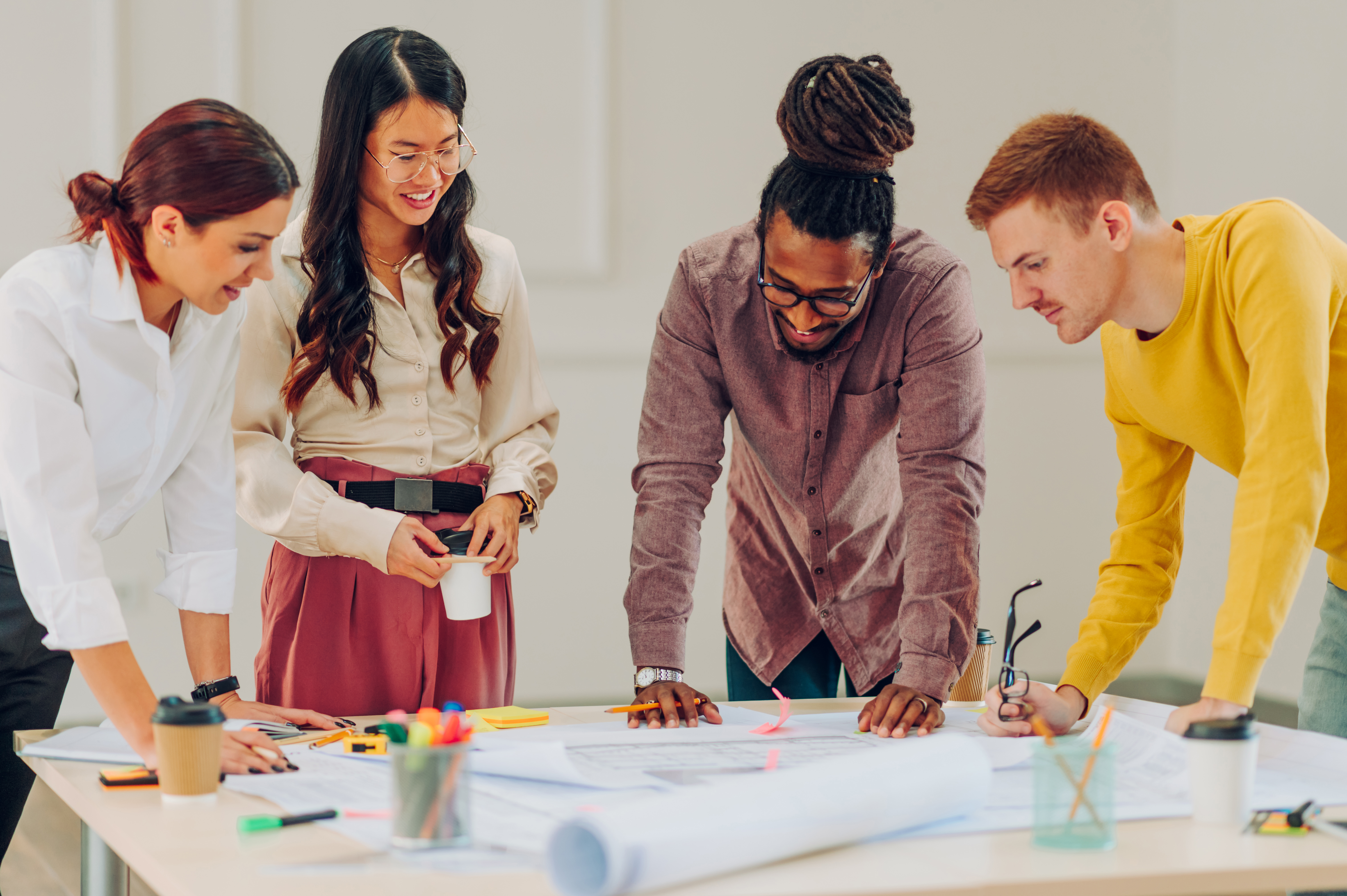 Grupo de trabalhadores composto por uma mulher de roupa branca e cabelo ruivo, uma mulher oriental de cabelos longos pretos, um homem negro de roupa social e cabelo afro com tranças e um homem branco de cabelos curtos e roupa amarela.