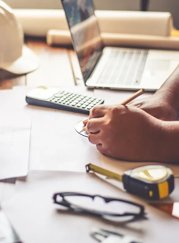 Homens em mesa de escritório com objetos de engenharia, calculadora, trena, capacete, papeis e computador