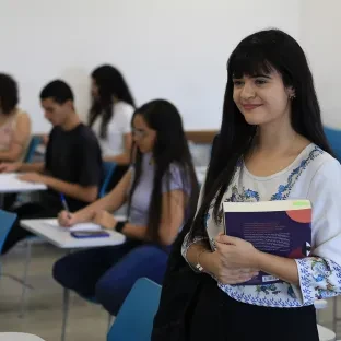 Mulher jovem de cabelo liso preto, em pé em sala de aula, segurando livros, com estudantes ao fundo da imagem.